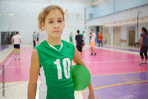 Girl in green poses with ball in gym during volleyball game, playing people out of focus