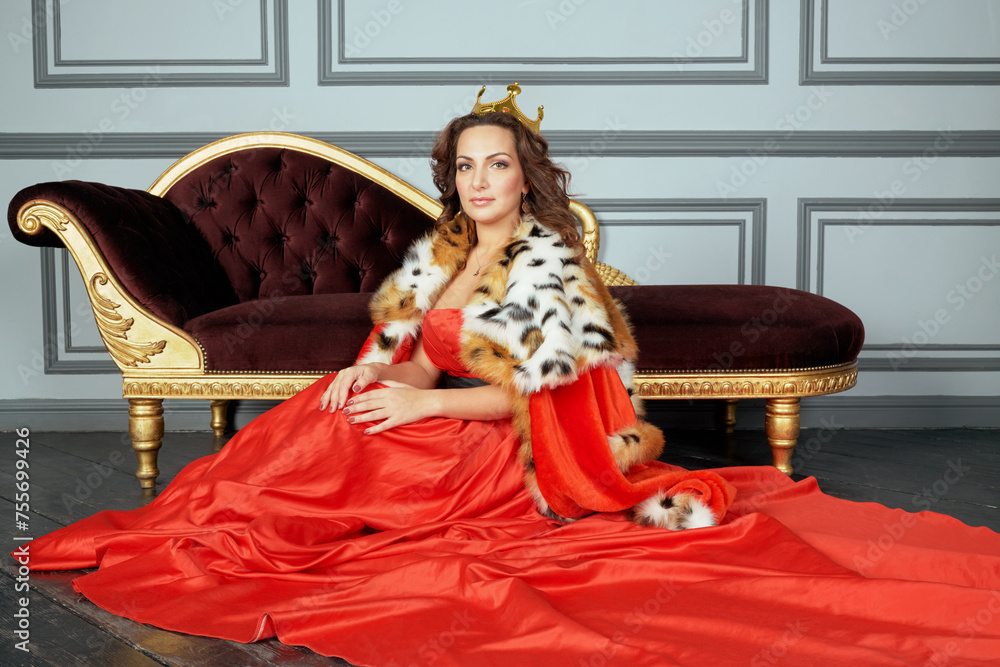 Fototapeta premium Woman in red dress, cloak and with crown on head sits on floor near couch in room