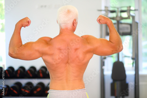 Middle-aged instructor shows his muscles in gym, back view, shallow dof