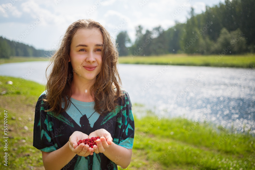 Happy girl teenager holds red wild strawberry under rain near river at summer day Happy girl teenager holds red wild strawberry under rain near river at summer day