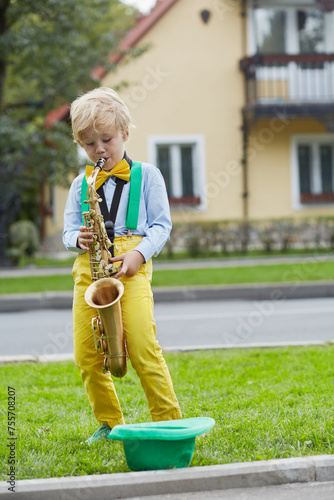 Little boy in dancing suit plays saxophone on grassy lawn against two-storied house near road, hat for earnings lies on grass photo