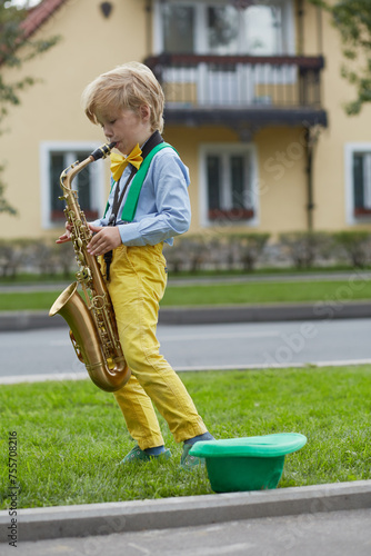 Little boy in dancing suit plays saxophone on grassy lawn against two-storied house near road, hat for earnings lies on grass photo