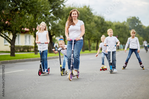 Woman and five children on scooters, roller skates and skateboard ride on street 