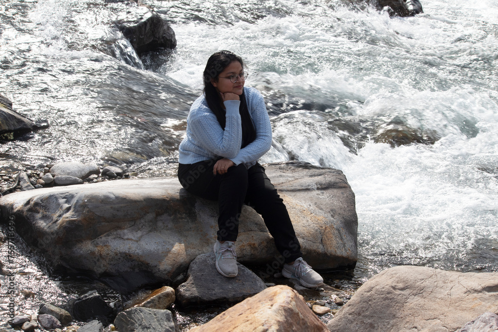 Smiling woman sits leisurely on an oversized rock, hand on chin, by the gushing waters of the Ganges River in Rishikesh, gazing contentedly at the scenic view, Uttarakhand Smiling woman sits leisurely on an oversized rock, hand on chin, by the gushing waters of the Ganges River in Rishikesh, gazing contentedly at the scenic view, Uttarakhand