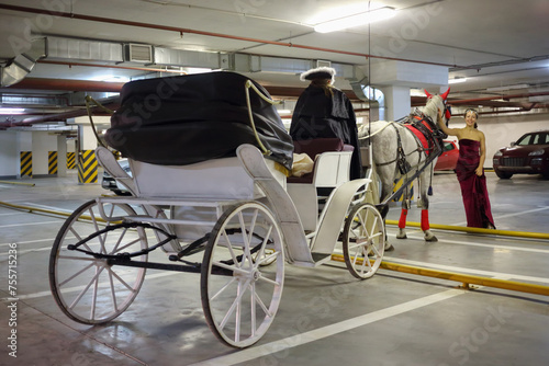 Woman stands near horse, coach and coachman in underground parking