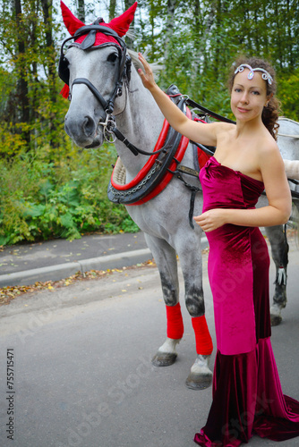 Pretty woman in red dress stands near horse in harness in green park