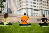 Man and two women practice yoga on grass near buildings at summer, back view