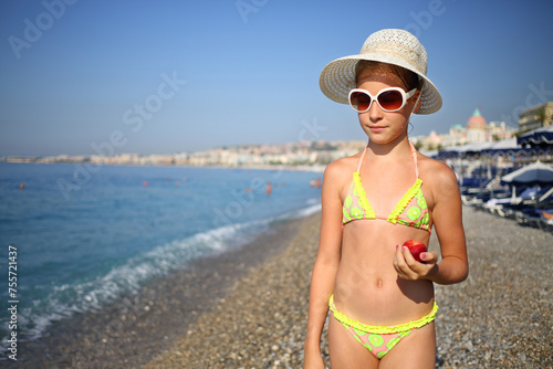Girl in hat and sunglasses holds red peach on beach on sunny day