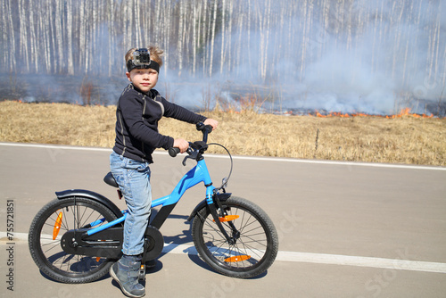 The little boy with the camera on his head with a bicycle standing on a road near a burning dry grass along the birch grove in the spring