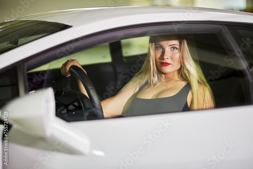 Young woman sits on driver seat of modern white car behind door glass at underground parking