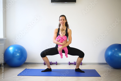 A smiling woman with a baby in the hands performing squats with wide-set legs in gym