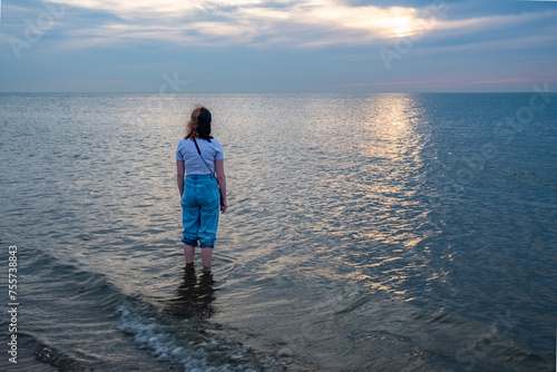 This poignant image captures a lone individual standing in the sea, gazing towards the distant horizon where the setting sun casts a silvery path across the water. The person's casual attire