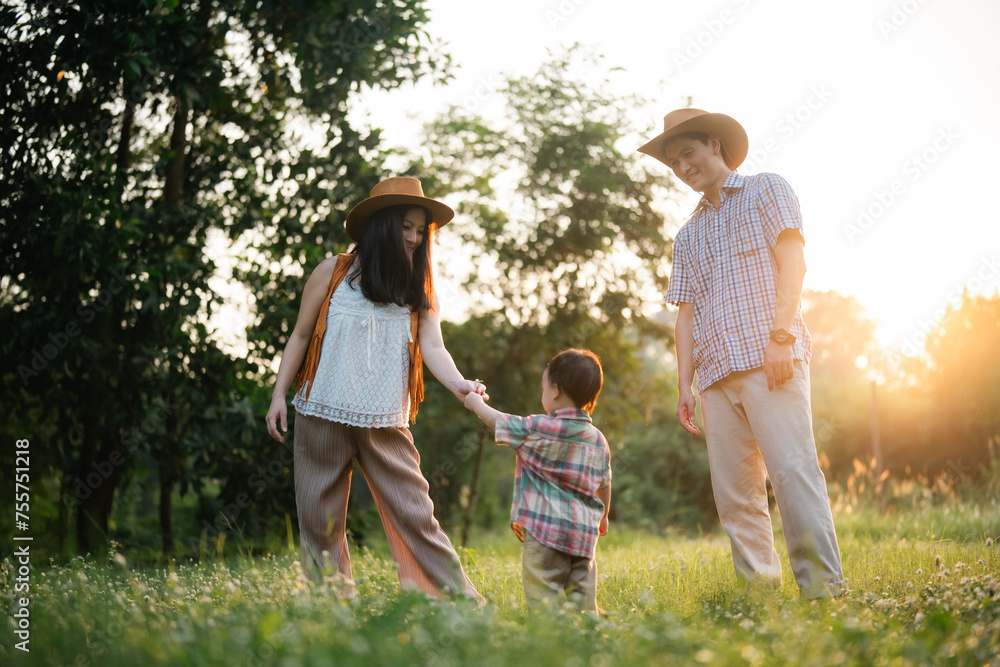 Obraz premium Asian family of three, a man, a woman and a child, are walking through a grassy field, summer activity in nature park is happy fun outdoor lifestyle