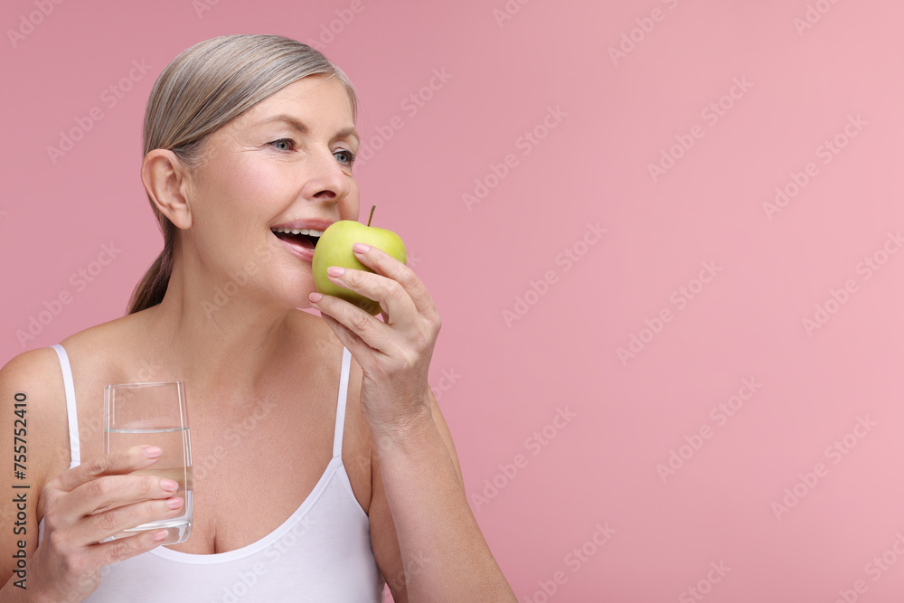 Beautiful woman with glass of water eating fresh apple on pink background, space for text. Vitamin rich food Beautiful woman with glass of water eating fresh apple on pink background, space for text. Vitamin rich food