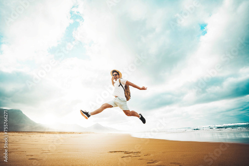 Happy traveler with hands up jumping at the beach - Delightful man enjoying success and freedom outdoors - Wanderlust, wellbeing, travel and summertime holidays concept 