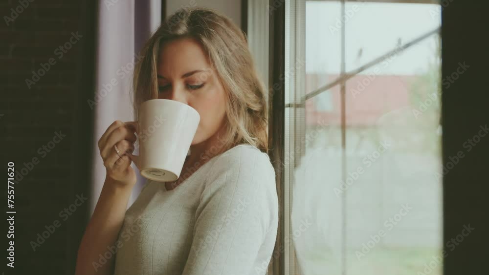 Blonde hair young woman holding cup of hot natural tea, coffee in hands drinking enjoys aroma of drink. Beauty face adult girl cozy room home near window full fog, cozy casual wear jeans t-shirt 4k Blonde hair young woman holding cup of hot natural tea, coffee in hands drinking enjoys aroma of drink. Beauty face adult girl cozy room home near window full fog, cozy casual wear jeans t-shirt 4k