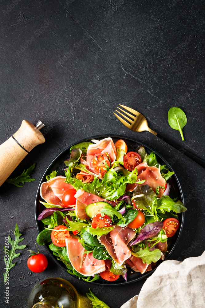 Green salad on black background. Fresh salad with jamon, green salad leaves and tomatoes. Top view with space for text.
