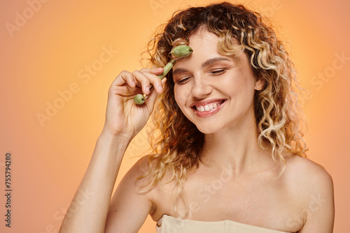 delighted woman with wavy hair and perfect face using jade roller on pink and yellow backdrop