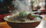 A steaming bowl of Vietnamese pho with thinly sliced meat, rice noodles, and bean sprouts. Fresh herbs, sliced chilies, and lime wedges are served on the side.