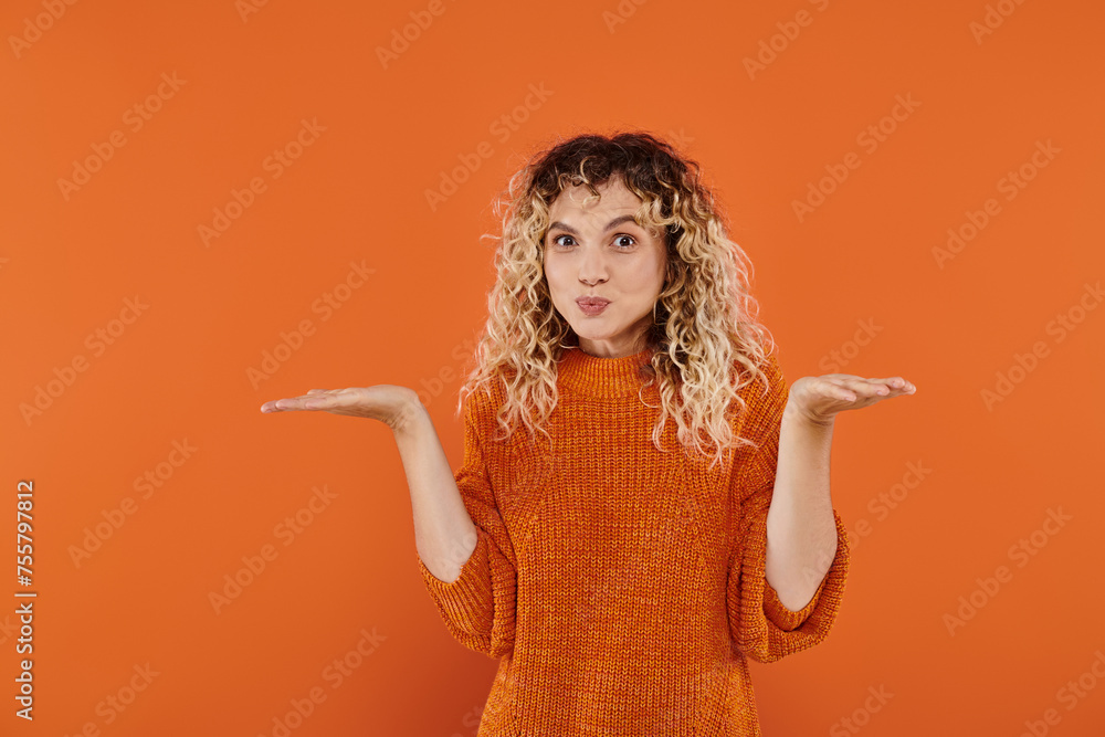 confused curly woman in knitted sweater puffing cheeks and showing shrug gesture on orange confused curly woman in knitted sweater puffing cheeks and showing shrug gesture on orange