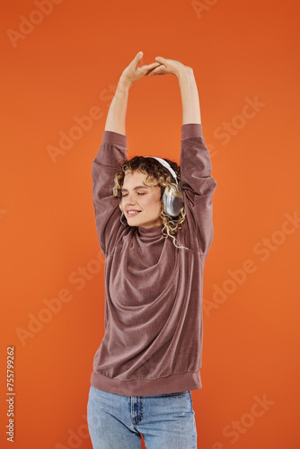 glad curly woman with closed eyes and raised hands listening music in wireless headphones on orange © LIGHTFIELD STUDIOS