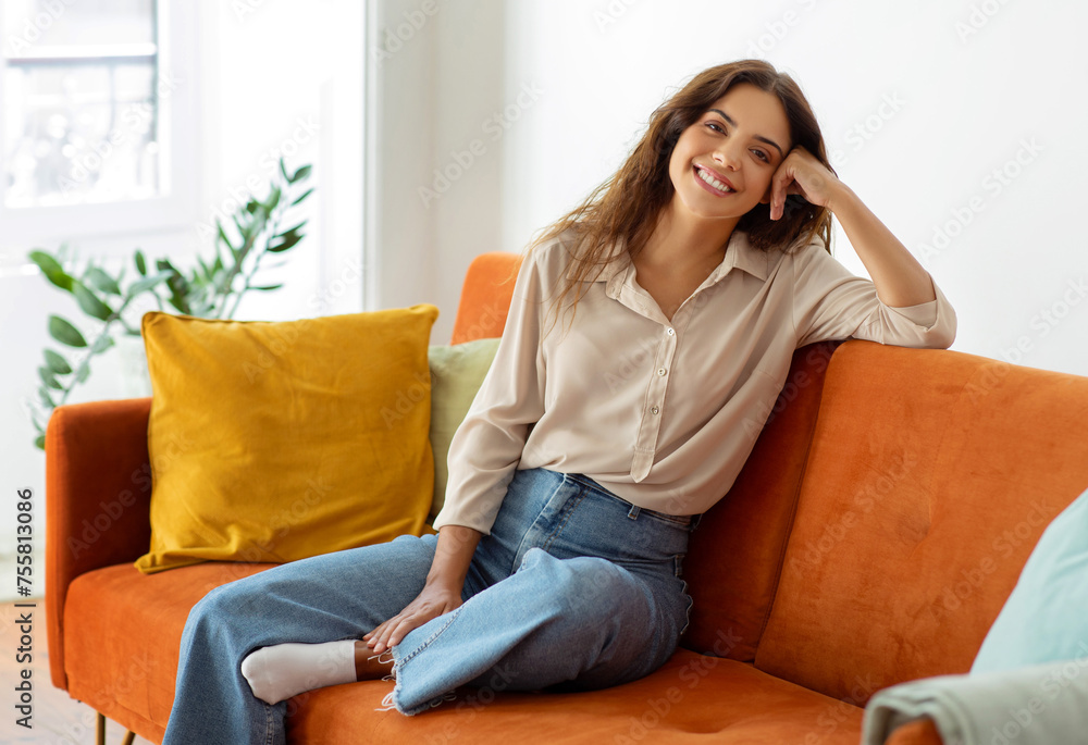 Cheerful young woman in casual clothes sitting comfortably on sofa at home