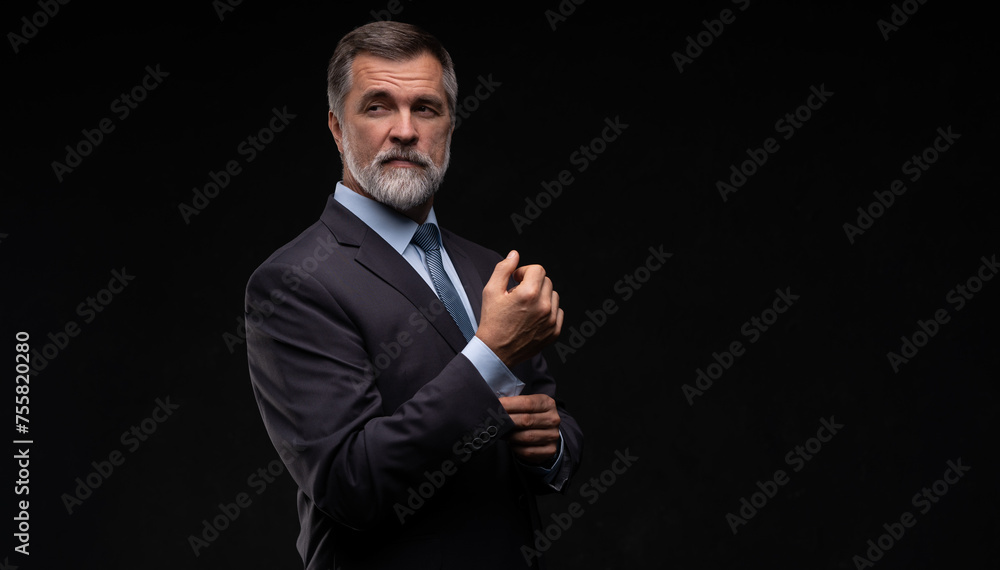 Portrait of a charming mature businessman dressed in suit posing while standing and looking at camera isolated over black background.