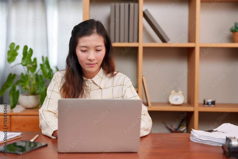 Asian adult female, engrossed in work, sitting at desk with laptop, tablet, papers; interior, daytime. Concentrated young office worker reviews documents, digital devices on wooden table, calm setting Asian adult female, engrossed in work, sitting at desk with laptop, tablet, papers; interior, daytime. Concentrated young office worker reviews documents, digital devices on wooden table, calm setting