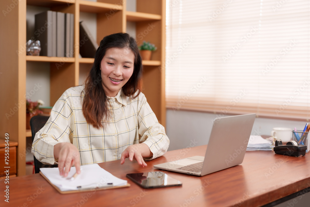 Cheerful Asian woman at wooden desk interacts with tablet, laptop beside, indoor plant backdrop. Happy professional engages with digital tablet, casual attire, work essentials arranged on desk. Cheerful Asian woman at wooden desk interacts with tablet, laptop beside, indoor plant backdrop. Happy professional engages with digital tablet, casual attire, work essentials arranged on desk.