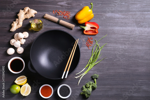Empty iron wok and chopsticks surrounded by ingredients on dark grey wooden table, flat lay. Space for text