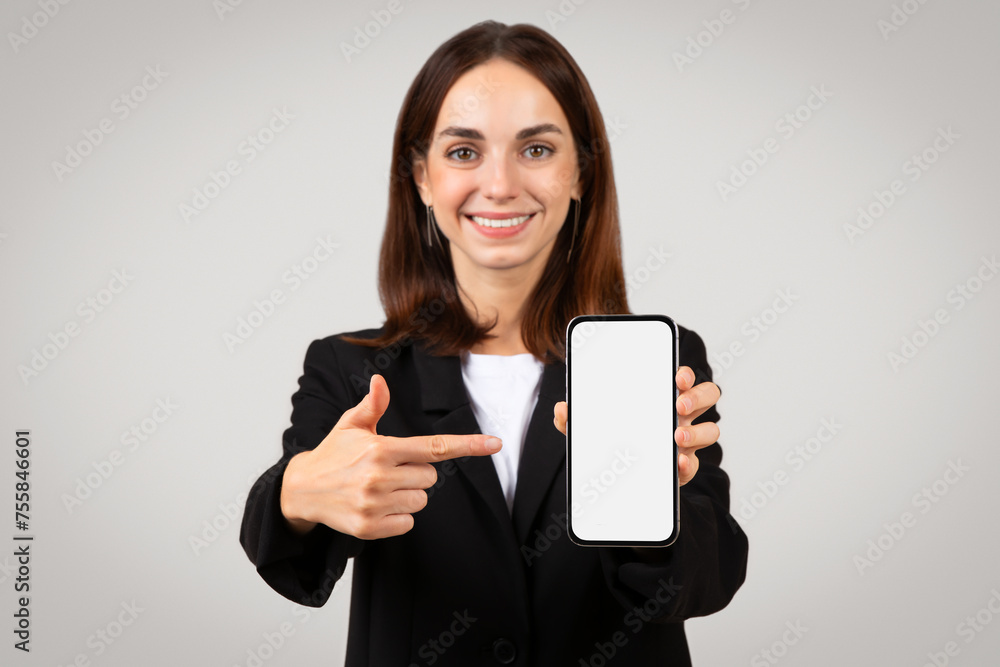 Friendly businesswoman presenting a smartphone with a blank screen and pointing at it Friendly businesswoman presenting a smartphone with a blank screen and pointing at it