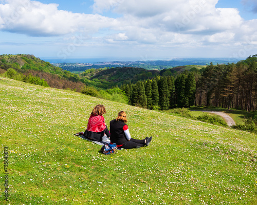 Basque country on border between France and Spain. Two women (unrecognizable  back view) having picnic at daisy covered meadow on mountain top  with beautiful view of  ocean and coastline near Hendaye © Elena Dijour