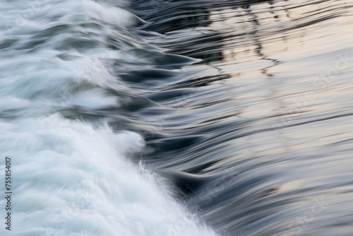 Rapid water flow over barrier with silky surface, river overflow over dam close up with splash and foam, abstract landscape pastel blue