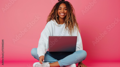 cheerful young woman sitting with her legs crossed, holding a laptop on her lap, wearing a white sweatshirt and ripped blue jeans, against a coral pink background