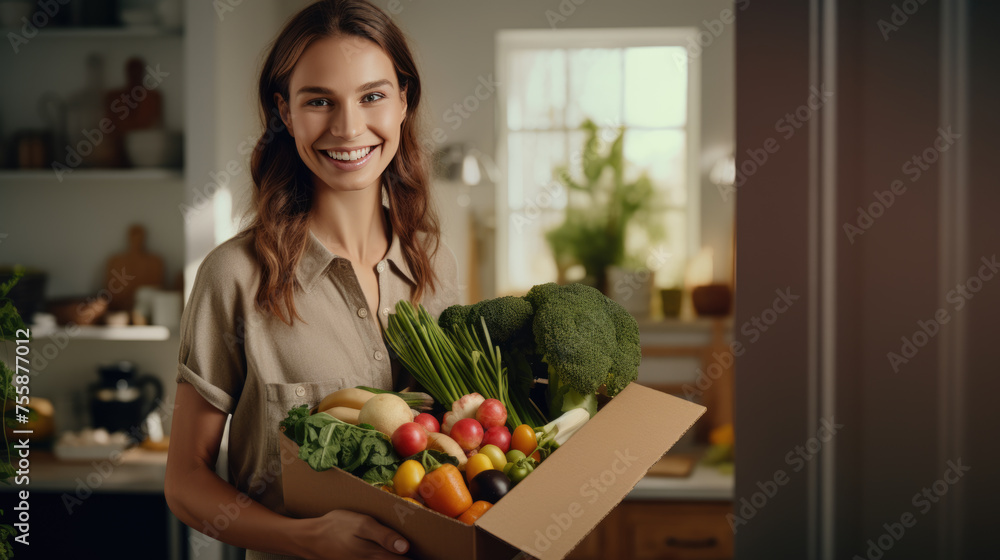 Smiling woman holding a box filled with fresh vegetables in a home kitchen setting.
