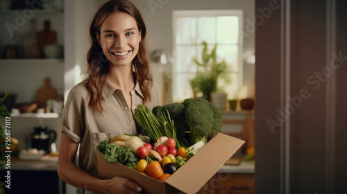 Smiling woman holding a box filled with fresh vegetables in a home kitchen setting. © MP Studio