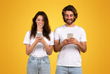A smiling woman and a man, both with dark hair and white t-shirts, are engrossed in their smartphones