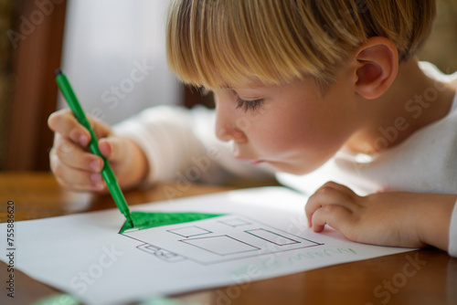 Small kid drawing a house on a table. Young caucasian boy sketching with markers at home on a sunny day. Blonde child coloring a sketch of a house with markers as his homework assignment.