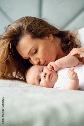 Happy Mother kissing her newborn baby. Closeup portrait of a loose haired loving mother kissing her child. Young mom with a beautiful infant child at home. Mum loves her infant son..
