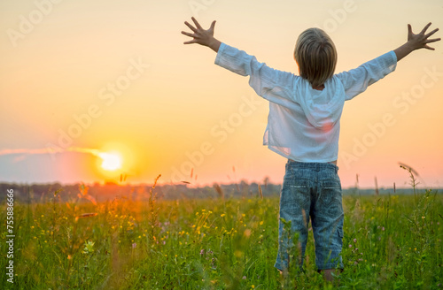 Happy boy with raised hands looks at the sunset  in summer. Boy in a field with his hands raised while sunrise. Freedom and relaxation concept. Blonde kid looking to the sun at the evening.