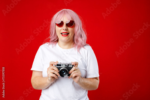 Portrait of a smiling young woman with pink hair standing with retro photo camera isolated over red background. Home hobby, lifestyle, travel, people concept