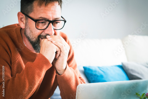 Focused young Caucasian man in glasses sit on sofa at home work online on laptop gadget. Concentrated boomer male typing on computer browse wireless internet on device. Technology concept. Business