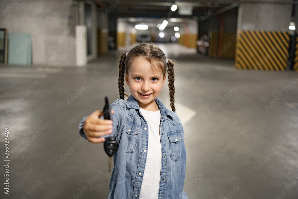 Portrait of cute little girl standing in underground parking lot in casual clothes holding keys opening car remotely, looking at camera. Happy child waiting for trip on weekend. Portrait of cute little girl standing in underground parking lot in casual clothes holding keys opening car remotely, looking at camera. Happy child waiting for trip on weekend.