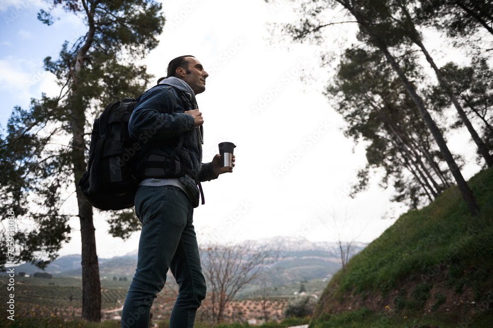 Side portrait of a young man with backpack and travel mug, during a trekking in the forest, mountains on the background Side portrait of a young man with backpack and travel mug, during a trekking in the forest, mountains on the background