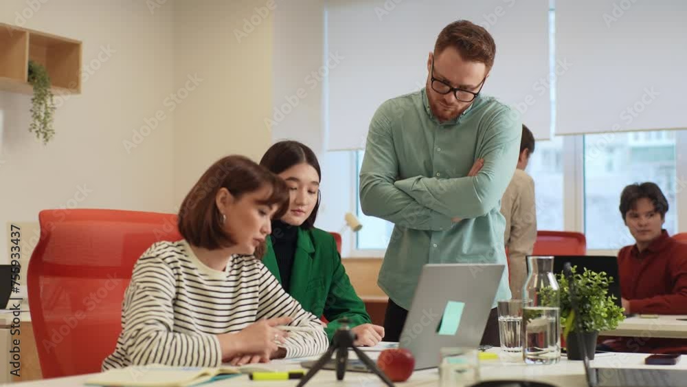 Skilled male team leader helping young female colleagues with online project on laptop computer, reviewing electronic document. Motivated millennial employees discussing development strategy with boss Skilled male team leader helping young female colleagues with online project on laptop computer, reviewing electronic document. Motivated millennial employees discussing development strategy with boss