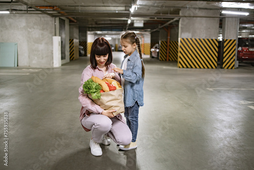 Caucasian mother with preschool daughter holding paper bag with vegetables in hands in underground parking lot. Happy family in casual clothes walking to their car after buying groceries for dinner.
