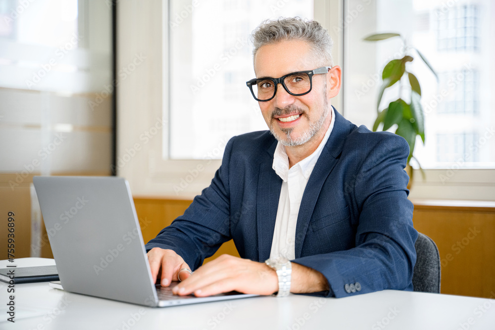 Friendly and positive mature male employee or manager using laptop, senior businessman in formal wear and glasses typing, responding to emails sitting in contemporary office Friendly and positive mature male employee or manager using laptop, senior businessman in formal wear and glasses typing, responding to emails sitting in contemporary office