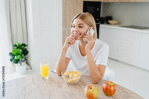 Cheerful caucasian young woman in casual clothes talking on the phone while having breakfast. Happy girl eating cereals and drinking orange juice at home kitchen