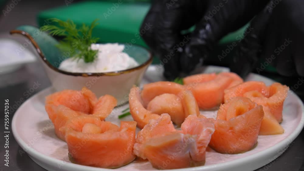 Close-up of marinated pieces of salmon on white plate with sauce and dill on table. Beautiful serving of pickled salmon. Hand of gloved chef turns dish and places it on table. Close-up of marinated pieces of salmon on white plate with sauce and dill on table. Beautiful serving of pickled salmon. Hand of gloved chef turns dish and places it on table.