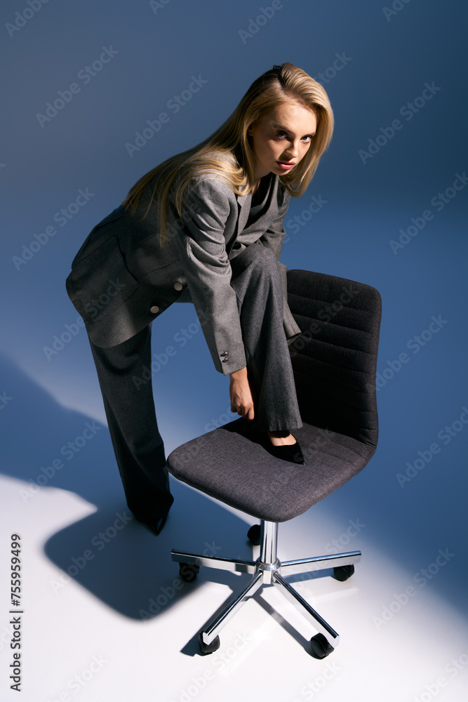 beautiful elegant woman in fashionable silver tuxedo posing near chair and looking at camera beautiful elegant woman in fashionable silver tuxedo posing near chair and looking at camera
